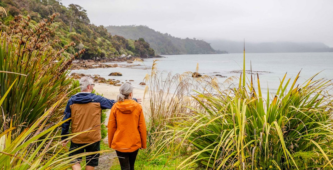 Couple walking along a coastal path on Stewart Island, surrounded by lush greenery and ocean views.
