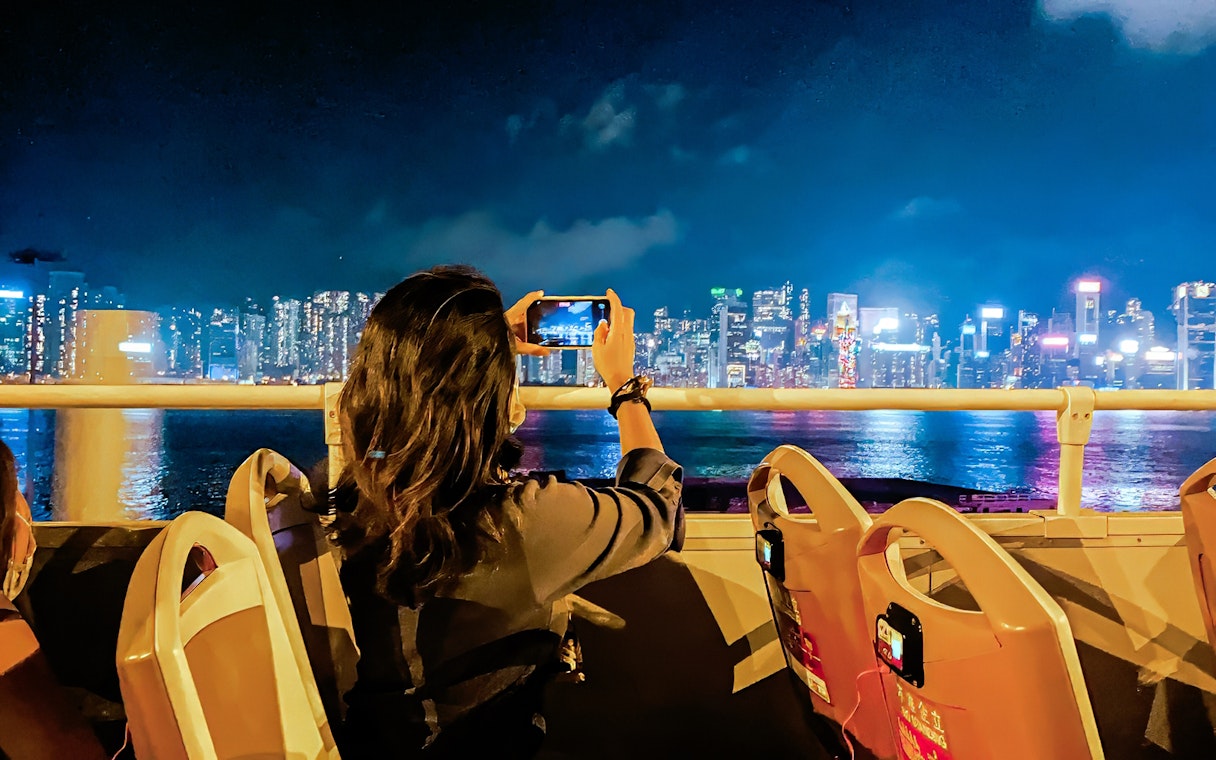 Person taking photo of Hong Kong skyline from open-top bus at night.