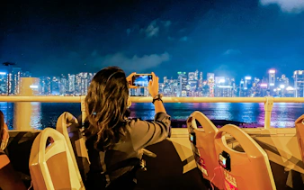 Person taking photo of Hong Kong skyline from open-top bus at night.