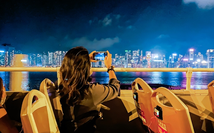 Person taking photo of Hong Kong skyline from open-top bus at night.