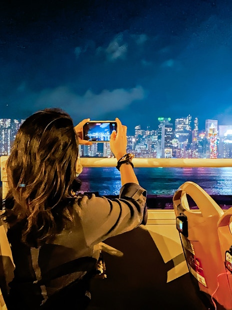 Person taking photo of Hong Kong skyline from open-top bus at night.