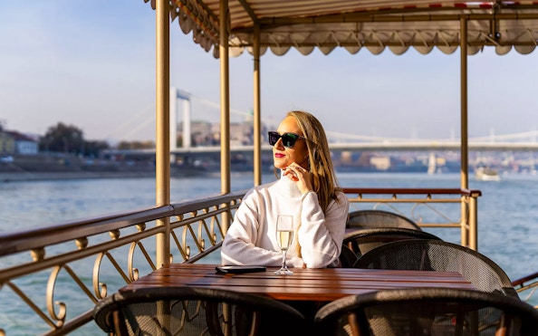 Woman enjoying a drink on a Budapest river cruise with bridge view.