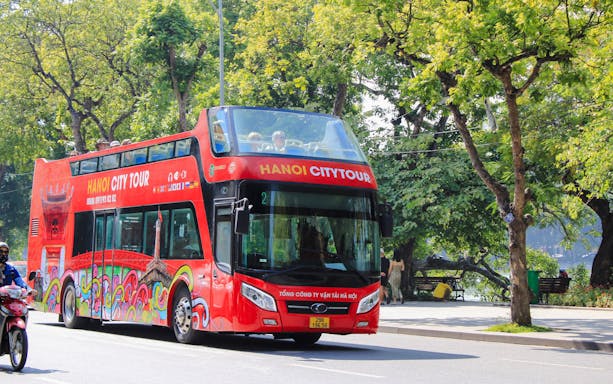 Hop on hop off tour bus driving through tree-lined street in Hanoi, Vietnam.