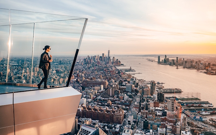 Person standing on The Edge observation deck overlooking New York City skyline at sunset.
