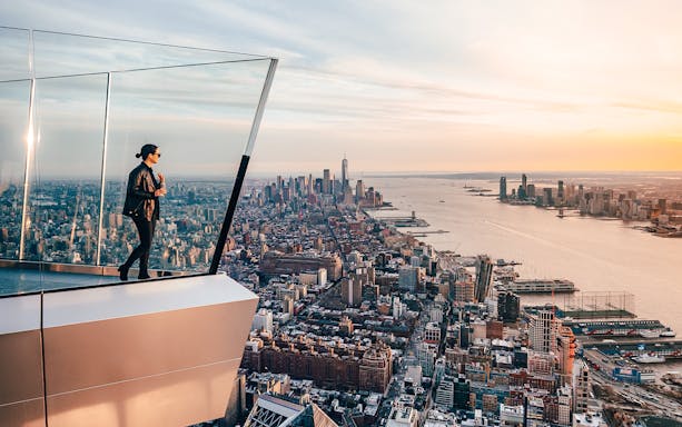Person standing on The Edge observation deck overlooking New York City skyline at sunset.