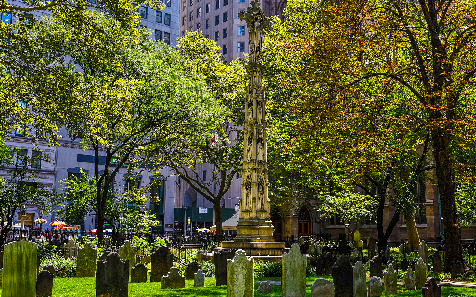 Trinity Churchyard gravestones and monument in Lower Manhattan, surrounded by trees and city buildings.