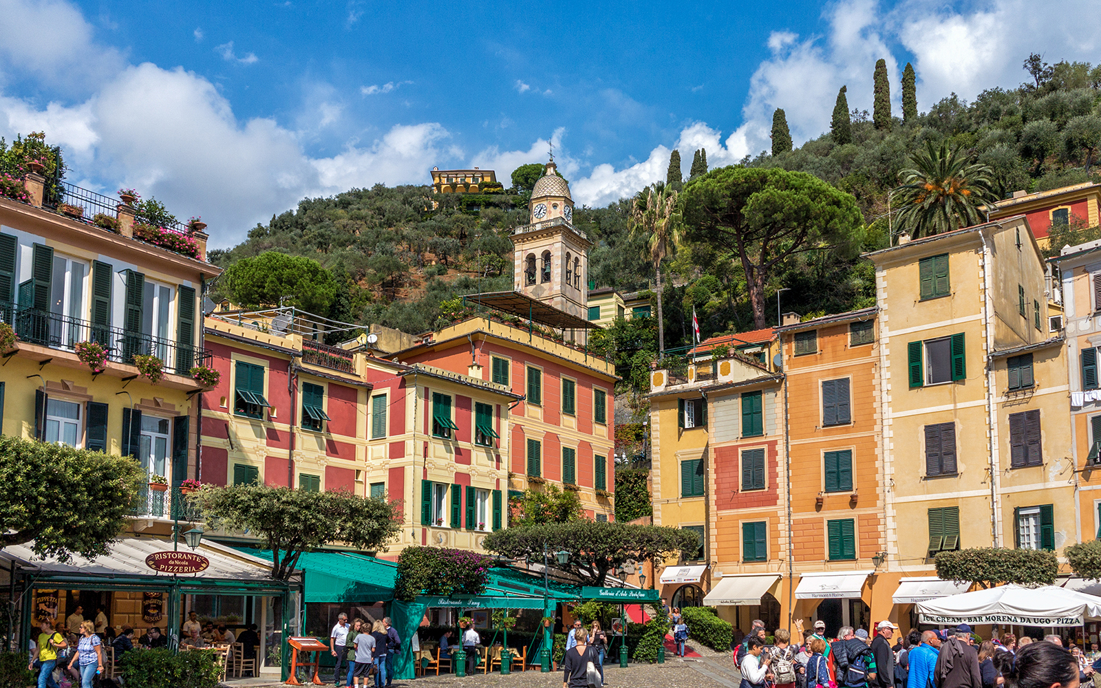 Colorful buildings and a clock tower in Portofino, Italy, with tourists exploring the area.
