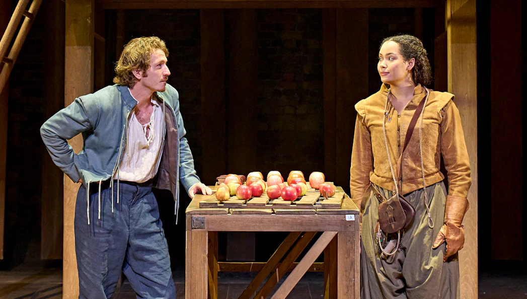 Two actors in period costumes perform a scene from "Hamnet" with a table of apples on stage.