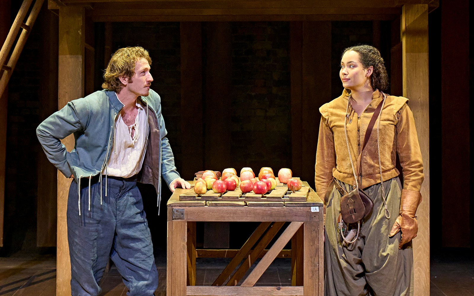 Two actors in period costumes perform a scene from "Hamnet" with a table of apples on stage.