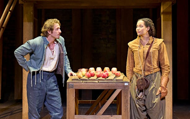 Two actors in period costumes perform a scene from "Hamnet" with a table of apples on stage.
