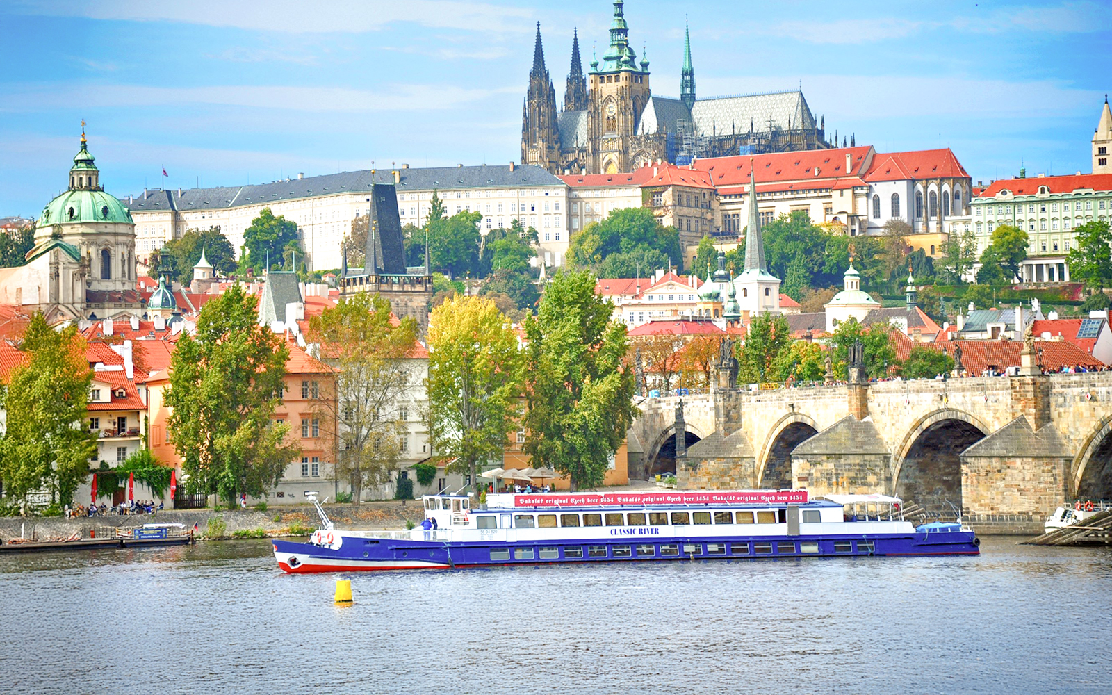 Cruise boat on Vltava River with Prague Castle and Charles Bridge in the background.