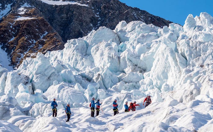 Group hiking on a glacier with ice formations and rocky mountain backdrop.
