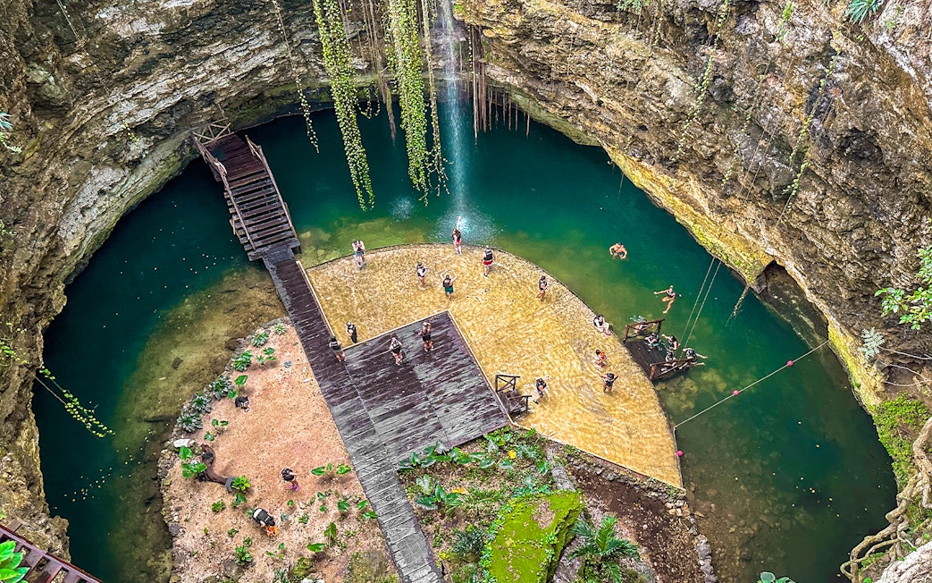 Tourists swimming in a natural Cenote pool surrounded by rocky cliffs and greenery.