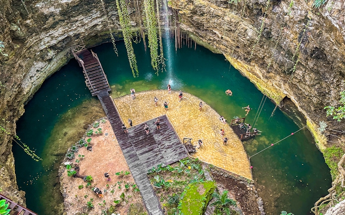 Tourists swimming in a natural Cenote pool surrounded by rocky cliffs and greenery.