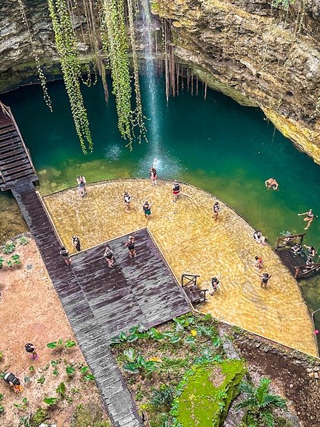 Tourists swimming in a natural Cenote pool surrounded by rocky cliffs and greenery.