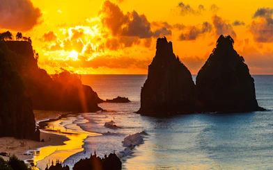 Sunset view of Dois Irmãos rocks at Noronha beach, Sao Paulo, with people on the shore.