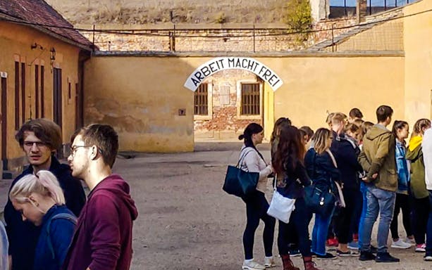 Guests exploring Terezin Concentration Camp courtyard under "Arbeit Macht Frei" sign.