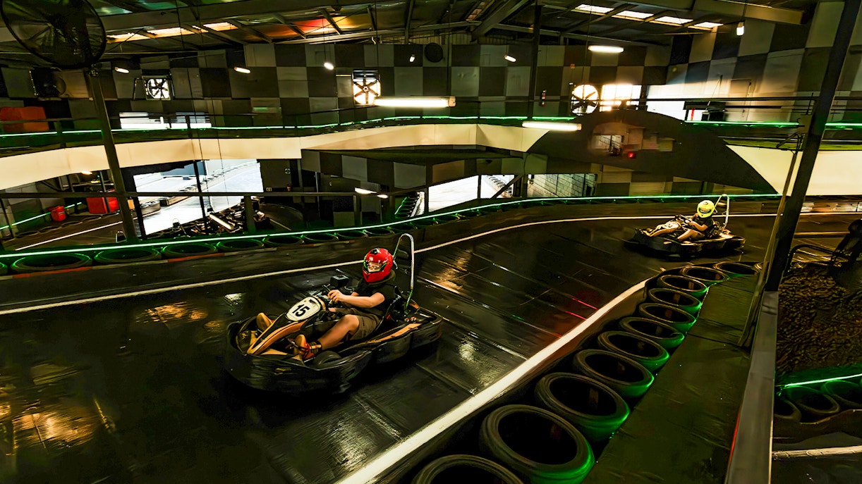 Indoor karting track with racers wearing helmets navigating a curve.