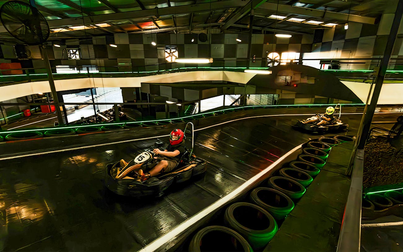 Indoor karting track with racers wearing helmets navigating a curve.