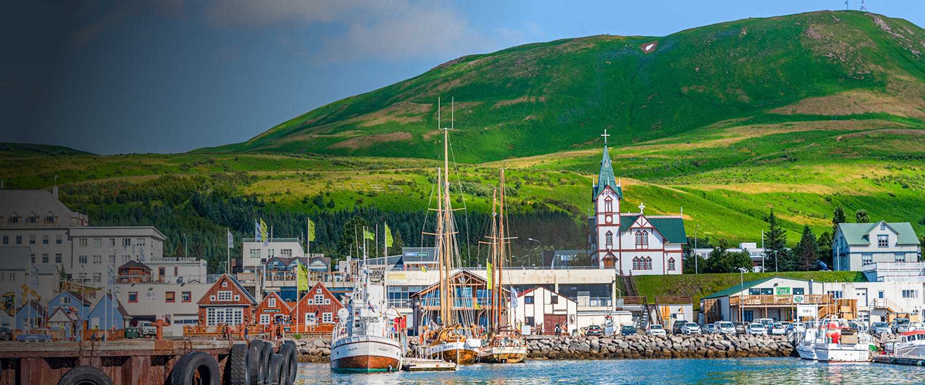 Fishing boats at Husavik port with colorful buildings and green hills, Iceland.