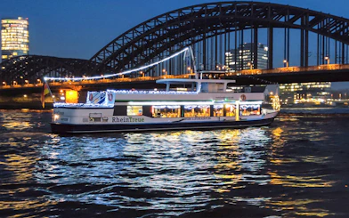 Cologne river cruise boat with festive lights near Hohenzollern Bridge at night.