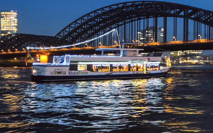 Cologne river cruise boat with festive lights near Hohenzollern Bridge at night.
