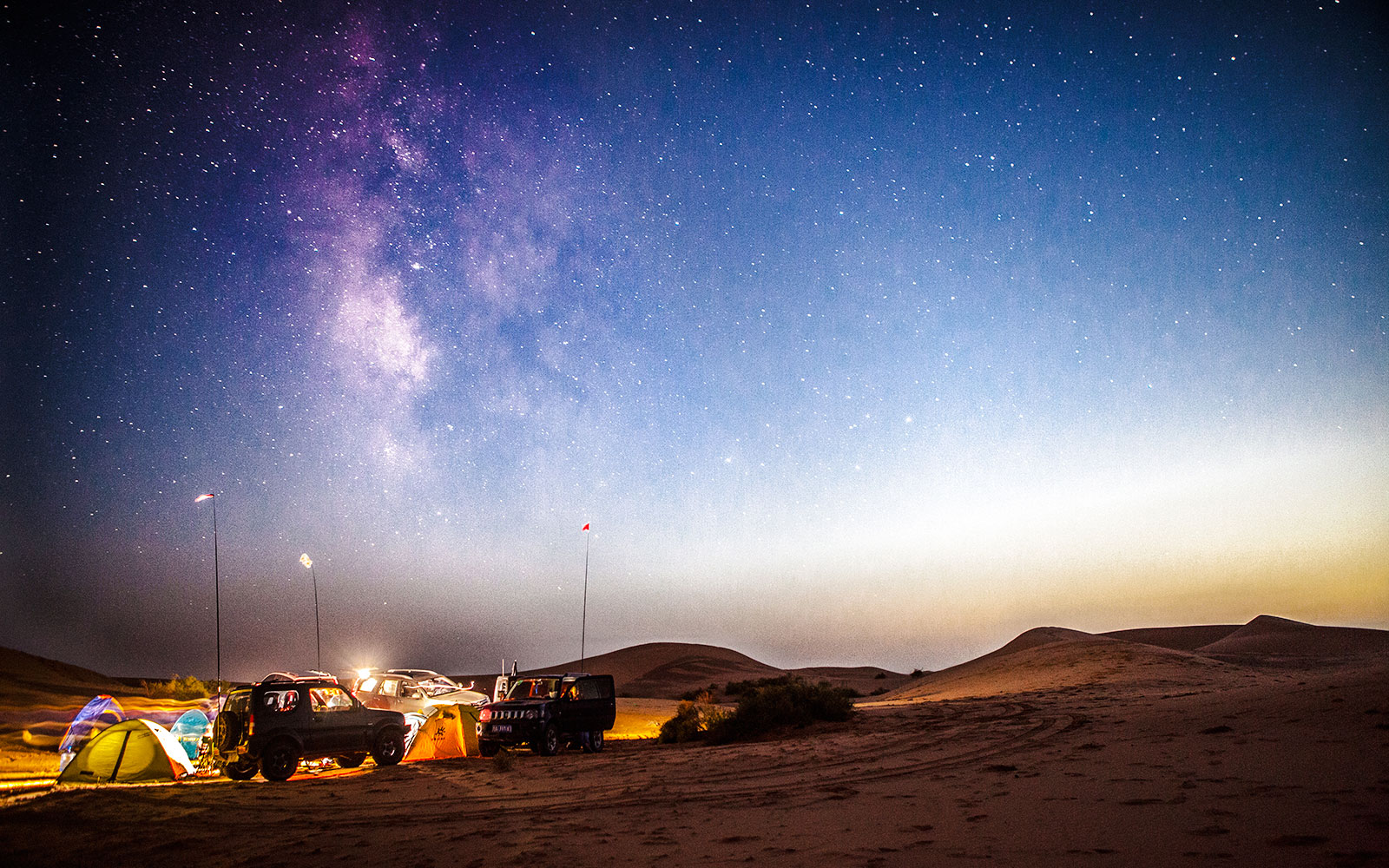 Milky Way rising over desert campsite at sunset with tents and vehicles.
