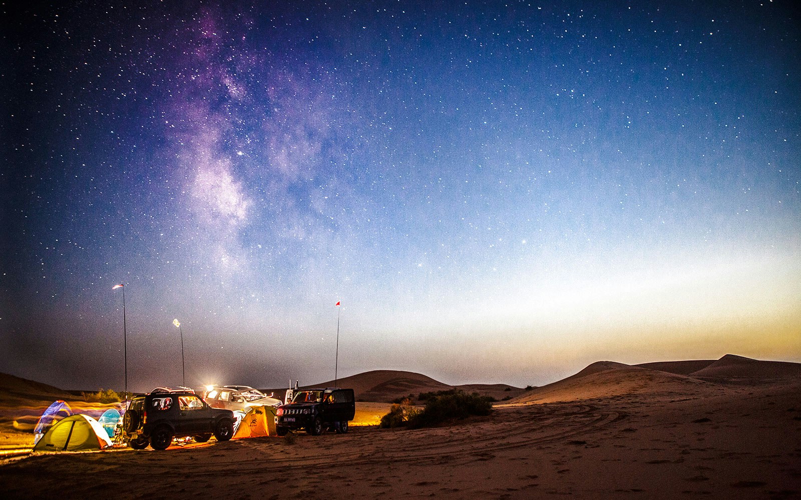 Milky Way rising over desert campsite at sunset with tents and vehicles.