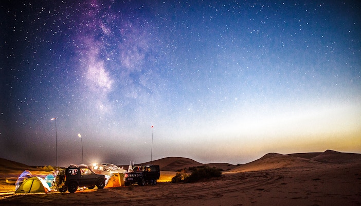 Milky Way rising over desert campsite at sunset with tents and vehicles.