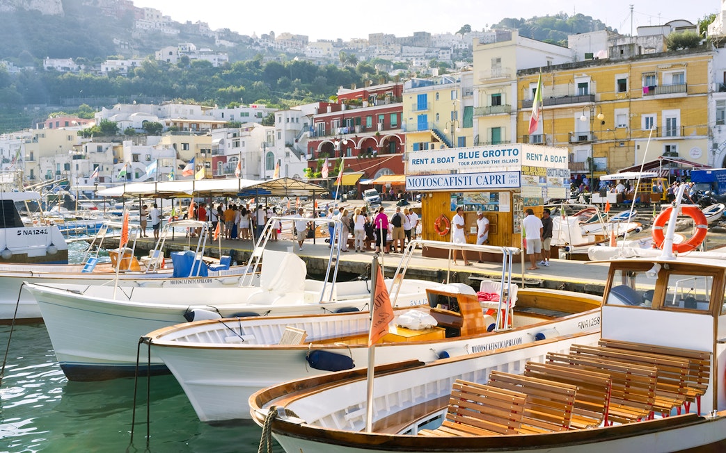 Boats docked at Capri Island harbor with tourists near Blue Grotto tour booth.