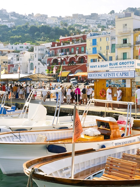 Boats docked at Capri Island harbor with tourists near Blue Grotto tour booth.