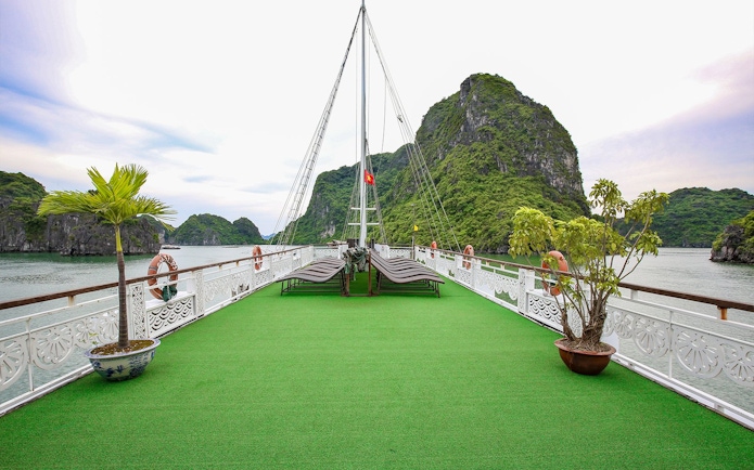 Cruise deck with lounge chairs and greenery, Halong Bay limestone cliffs in background.