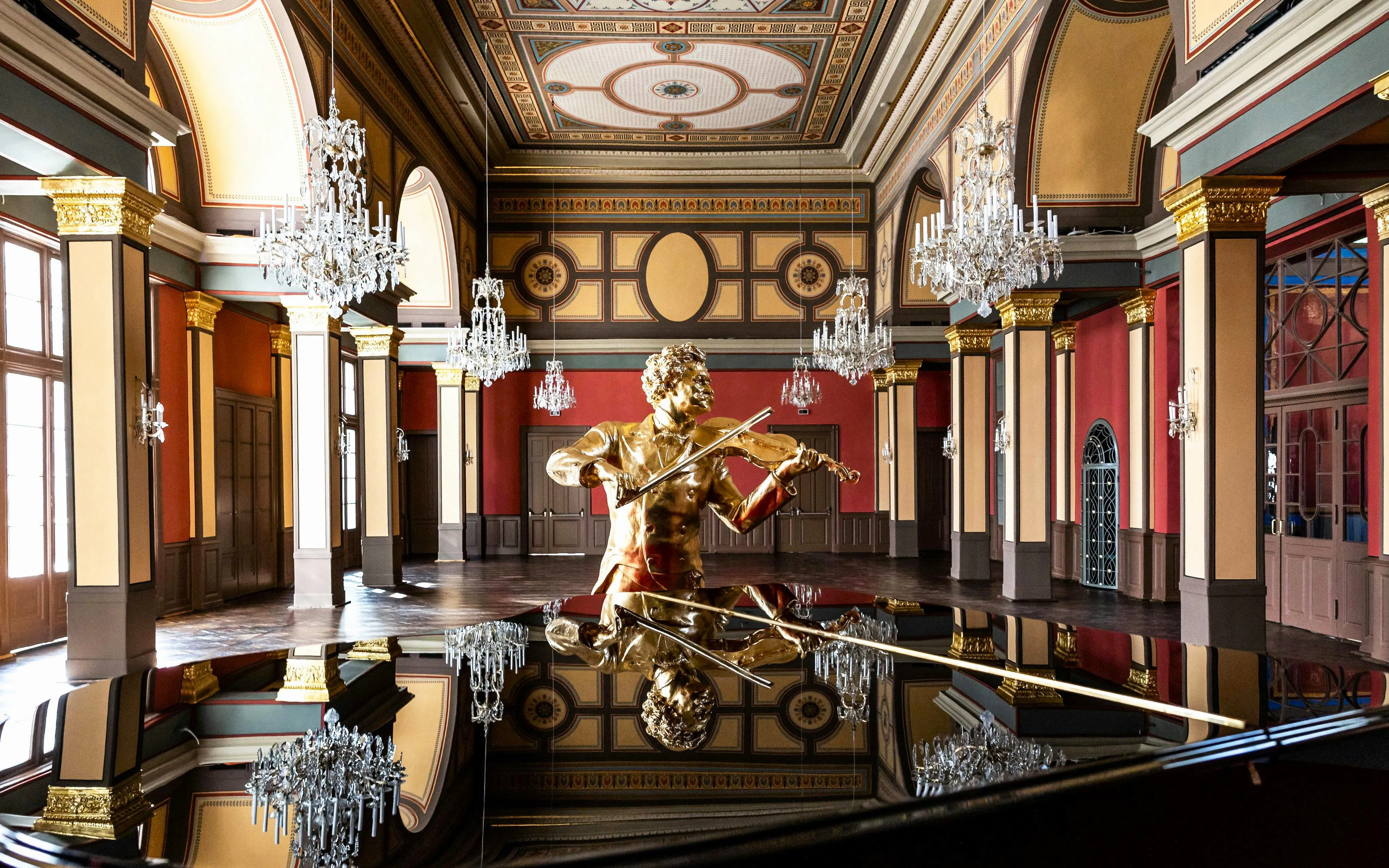 Golden violinist statue in ornate Vienna concert hall with chandeliers.