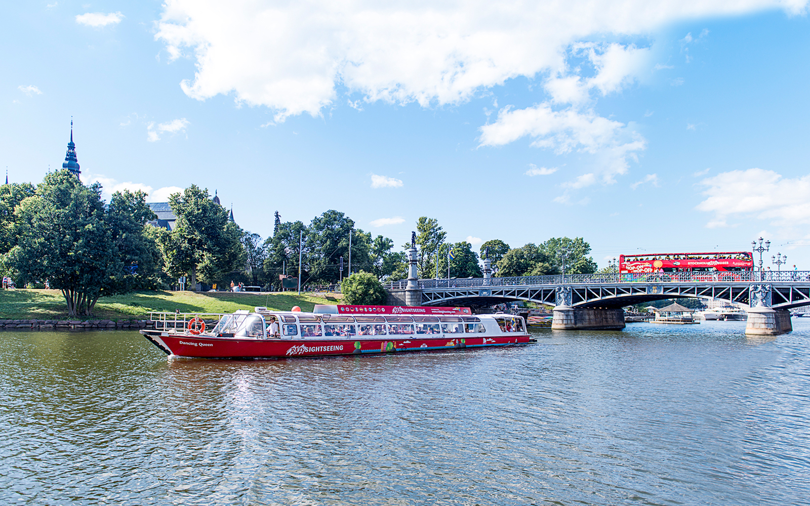Red sightseeing boat on Stockholm's waterway near a bridge and greenery.