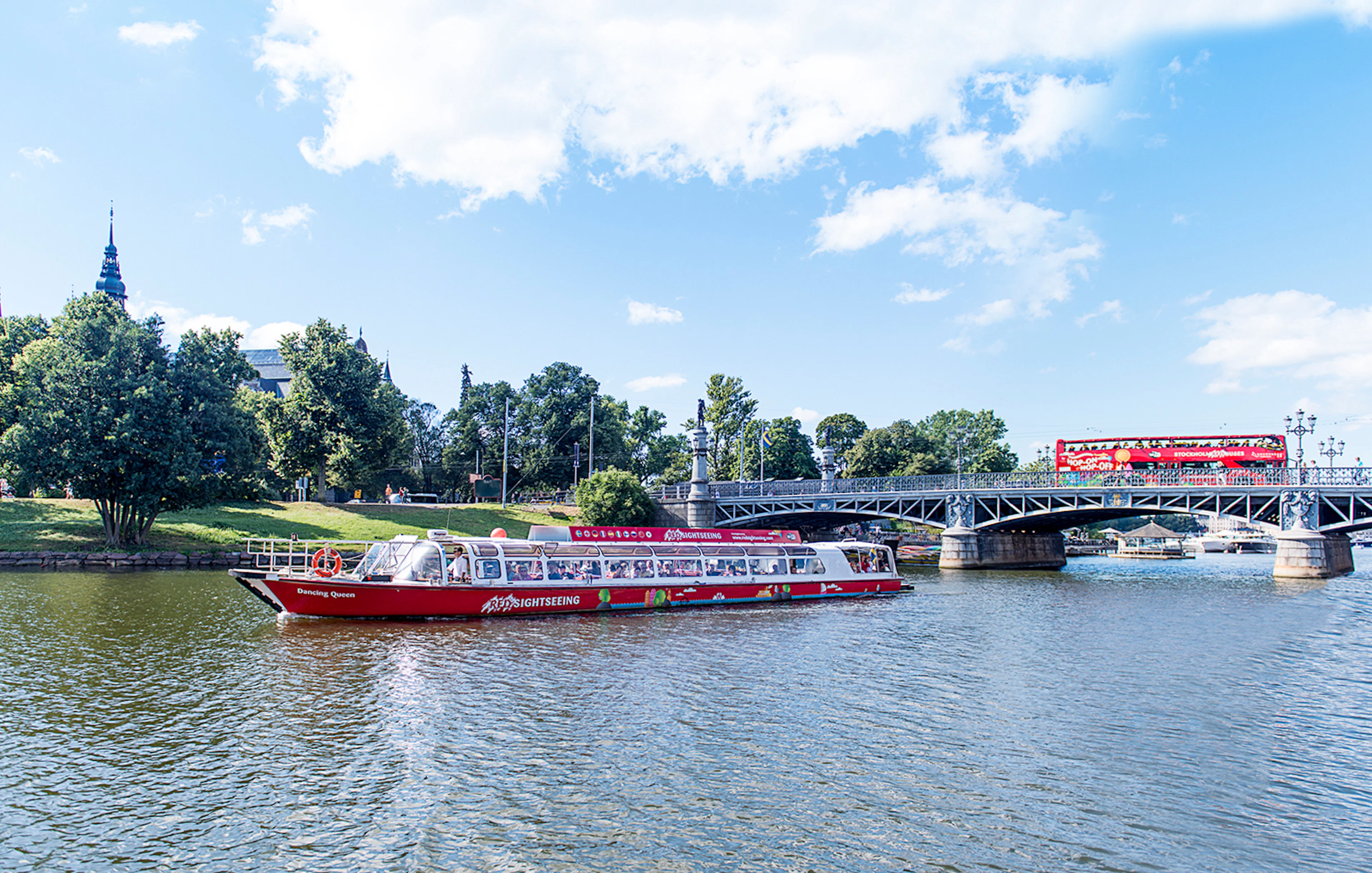 Red sightseeing boat on Stockholm's waterway near a bridge and greenery.