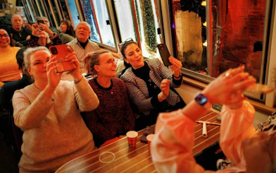 People enjoying a canal cruise during the Amsterdam Light Festival, capturing the view on their phones.