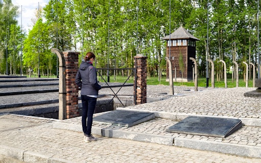 Visitor at Auschwitz Birkenau memorial site near watch tower and plaques.