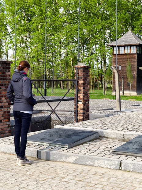 Visitor at Auschwitz Birkenau memorial site near watch tower and plaques.