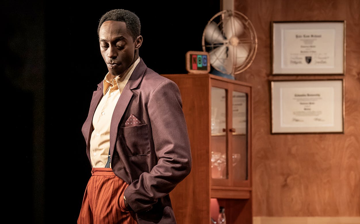 Man in vintage suit standing in retro-themed room with diplomas and fan.