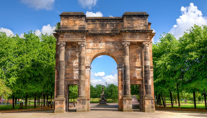 McLennan Arch in Glasgow Green, Scotland, surrounded by trees under a blue sky.