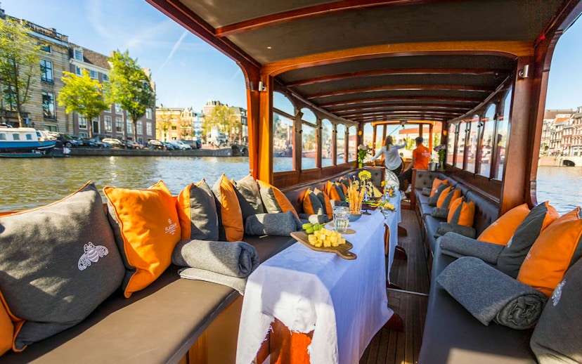Canal boat in Amsterdam with wine and cheese setup, city buildings in background.