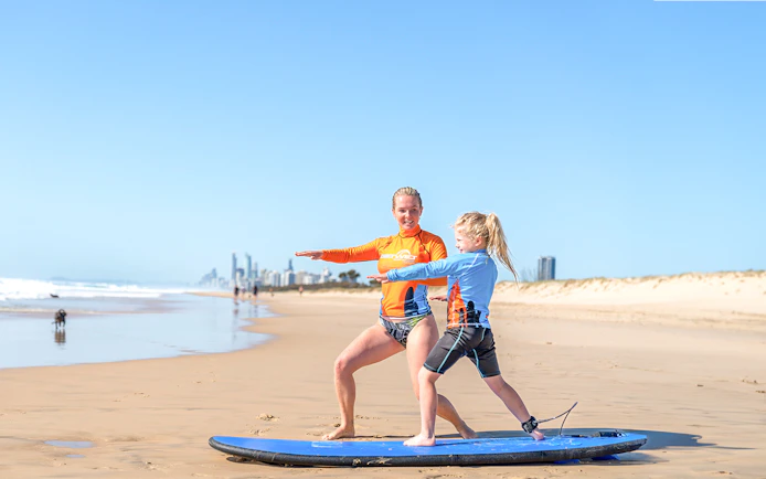Surf instructor teaching a child on a beach with city skyline in the background.