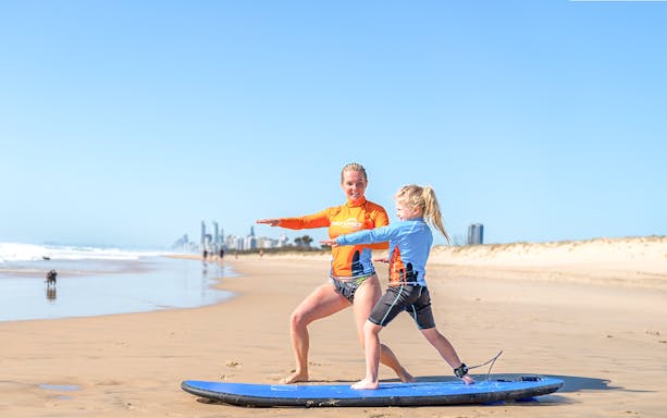 Surf instructor teaching a child on a beach with city skyline in the background.