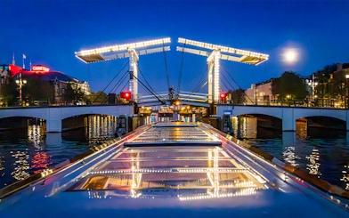 Boat approaching illuminated Skinny Bridge in Amsterdam at night.