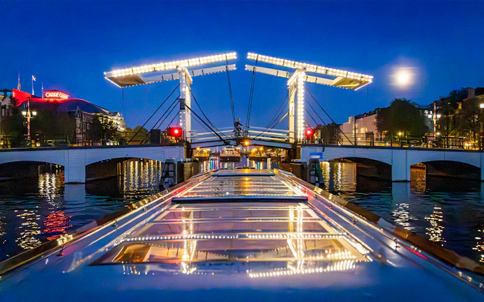 Boat approaching illuminated Skinny Bridge in Amsterdam at night.