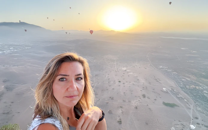 Woman taking selfie in hot air balloon over Marrakech desert at sunrise.