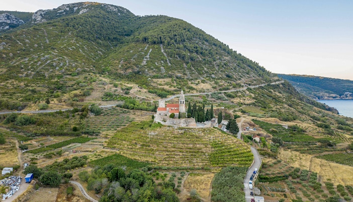 Aerial view of St. Nicolas Church on Mount Hum in Komiza, surrounded by terraced fields and hills.