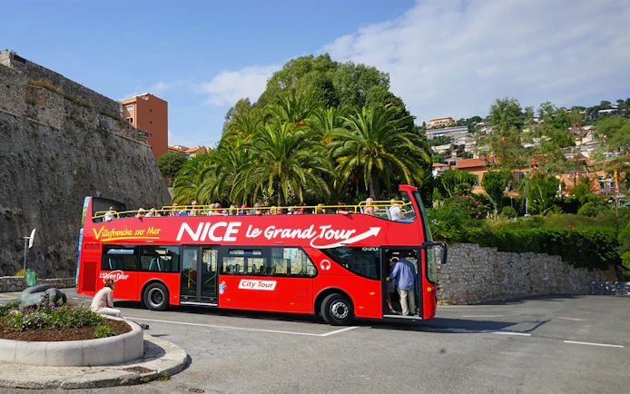 Red double-decker bus on Nice & Villefranche hop-on hop-off tour with palm trees in background.
