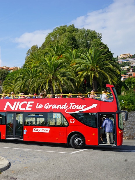 Red double-decker bus on Nice & Villefranche hop-on hop-off tour with palm trees in background.