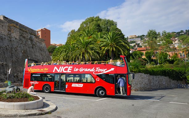 Red double-decker bus on Nice & Villefranche hop-on hop-off tour with palm trees in background.